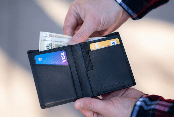 A person holding a black wallet with visible cash and two credit cards.