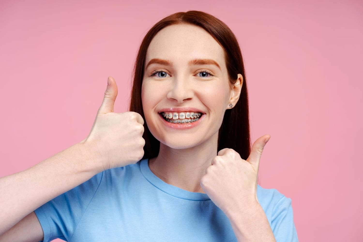 A smiling person with braces from Best Orthodontics gives two thumbs up, proudly wearing a light blue shirt against a vibrant pink background.