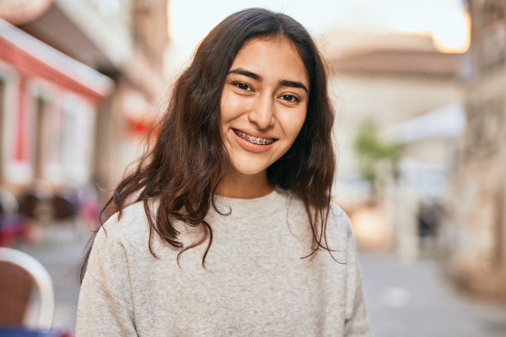 A young woman with long dark hair and braces, courtesy of Orthoone Orthodontics, smiles while standing on an outdoor street with blurred buildings in the background.