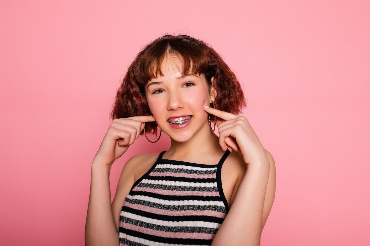 Young person with braces smiling, pointing to their cheeks, on a pink background.