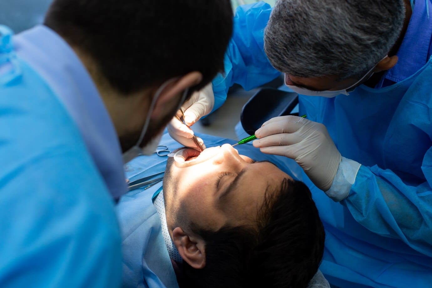 Two dentists in blue scrubs perform a dental procedure on a patient lying on an examination chair.