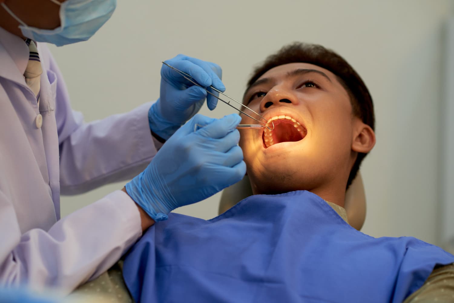 Dentist in protective gear examines a patient's open mouth with dental tools