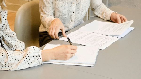Two people sit at a table reviewing documents, with one person taking notes in a notebook.