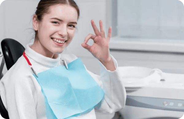 A person in a dental chair wearing a bib smiles and makes an OK gesture.