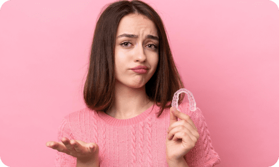 A woman in a pink sweater holds a clear dental aligner in one hand, while gesturing with the other against a pink background.