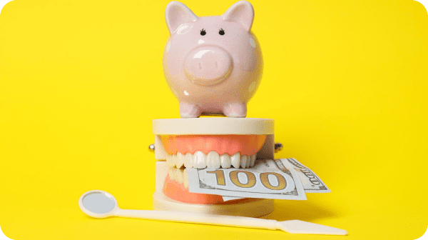 A piggy bank sits atop a model mouth holding a $100 bill, with a dental mirror and tool nearby, all against a yellow background.