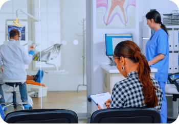 A woman fills out a form in a waiting area while a dentist attends to a patient in the examination room. A nurse works at a computer nearby.