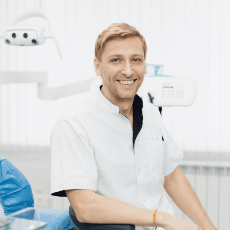 A person in a white coat smiles while seated in a dental office with equipment and a dental chair in the background.