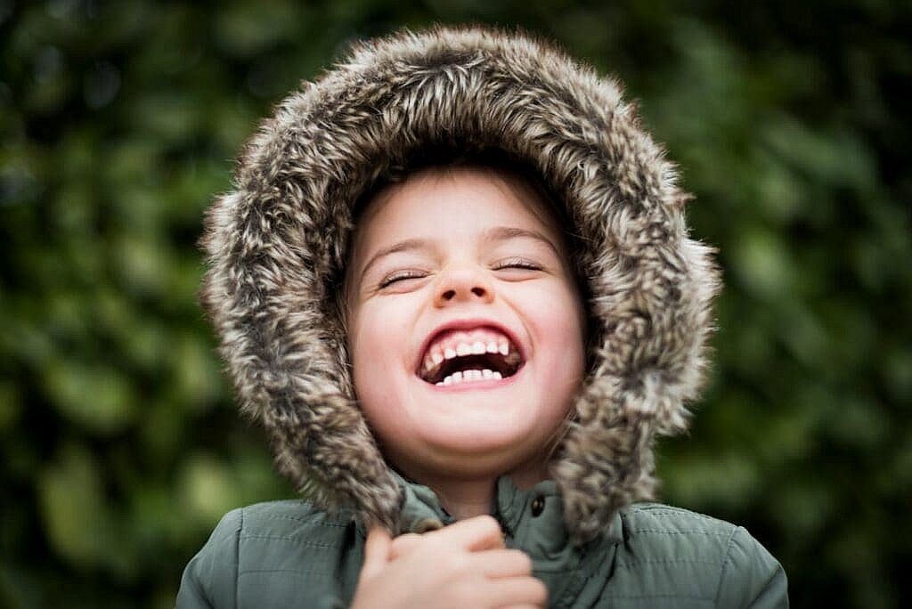 A child in a fur-lined hooded coat, adorned with affordable braces, laughs joyfully outdoors with a green foliage background in Austin or Houston, TX